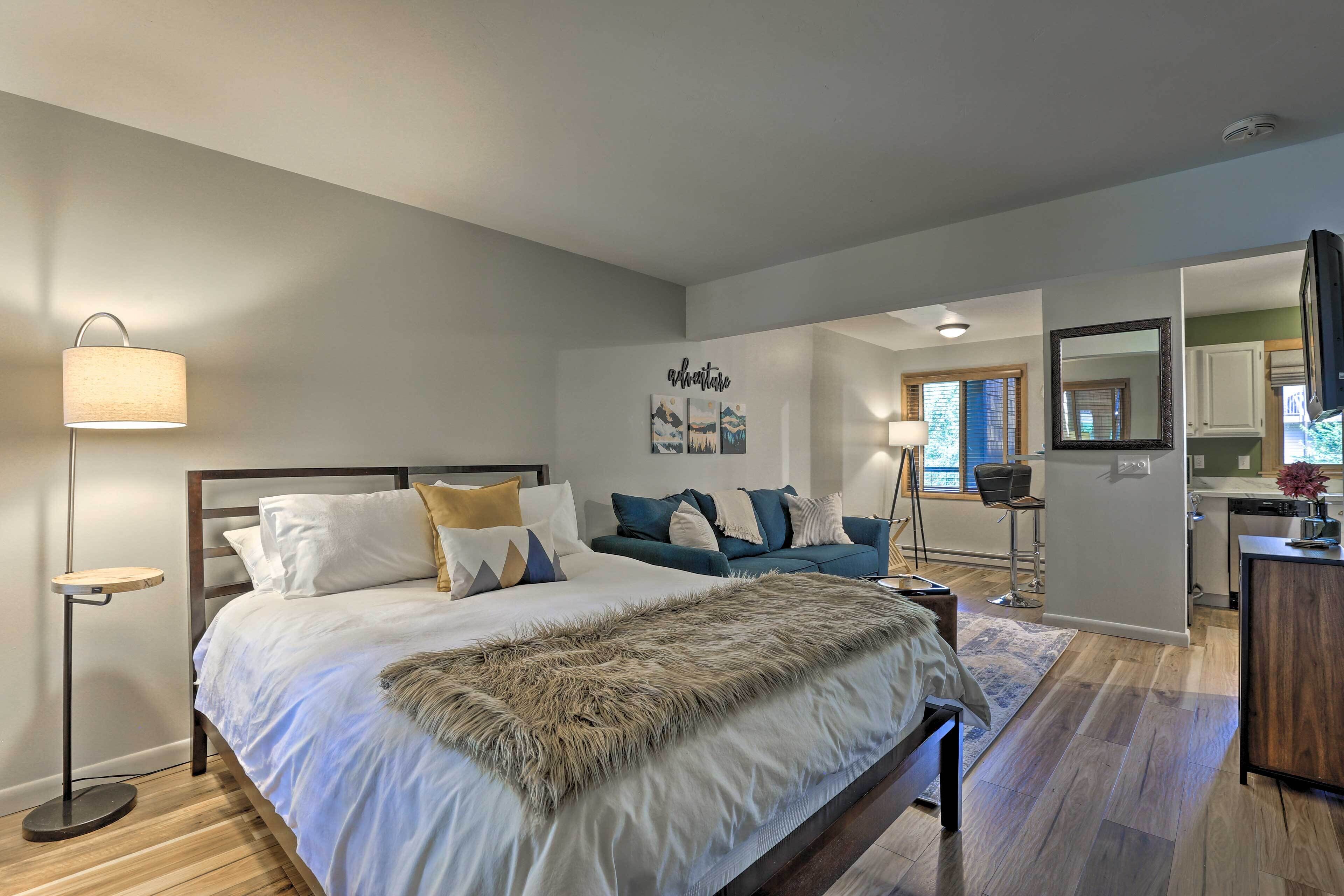 Une chambre moderne avec un grand lit, une lampe sur pied, un canapé et un coin cuisine visible à travers une porte ouverte.