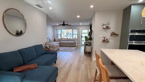 A modern living room with a blue sofa, a dining area with a white marble table, and a kitchen with built-in appliances.