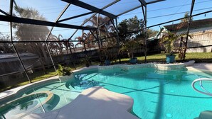 A clear, blue swimming pool with a curved design, surrounded by a fenced area with palm trees and a wooden fence in the background.