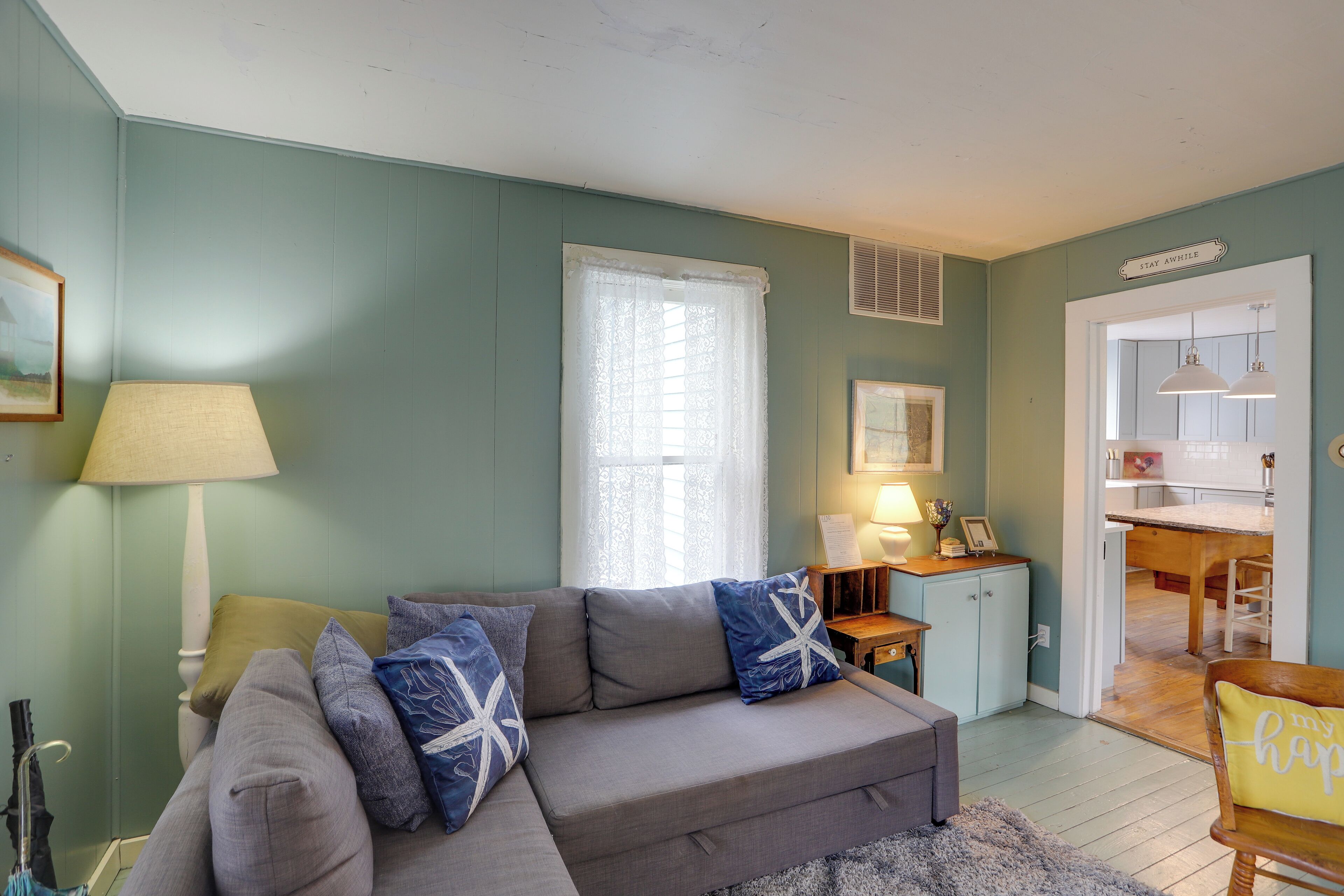 A living room with a grey sofa, a lamp, and a window with lace curtains.