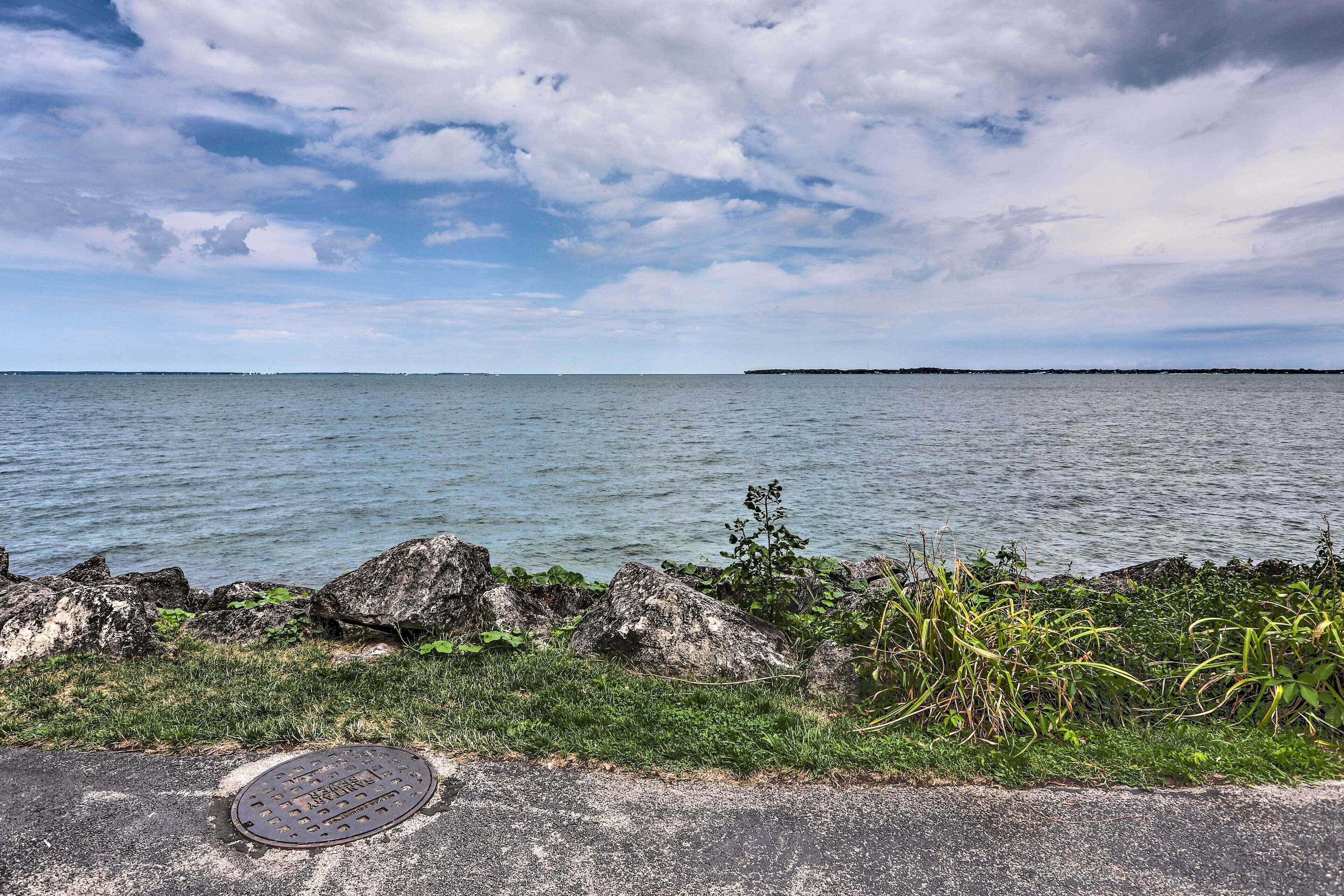 Una strada costiera con un coperchio in metallo per pozzetto, una zona erbosa con rocce e vista sul mare sotto un cielo parzialmente nuvoloso.