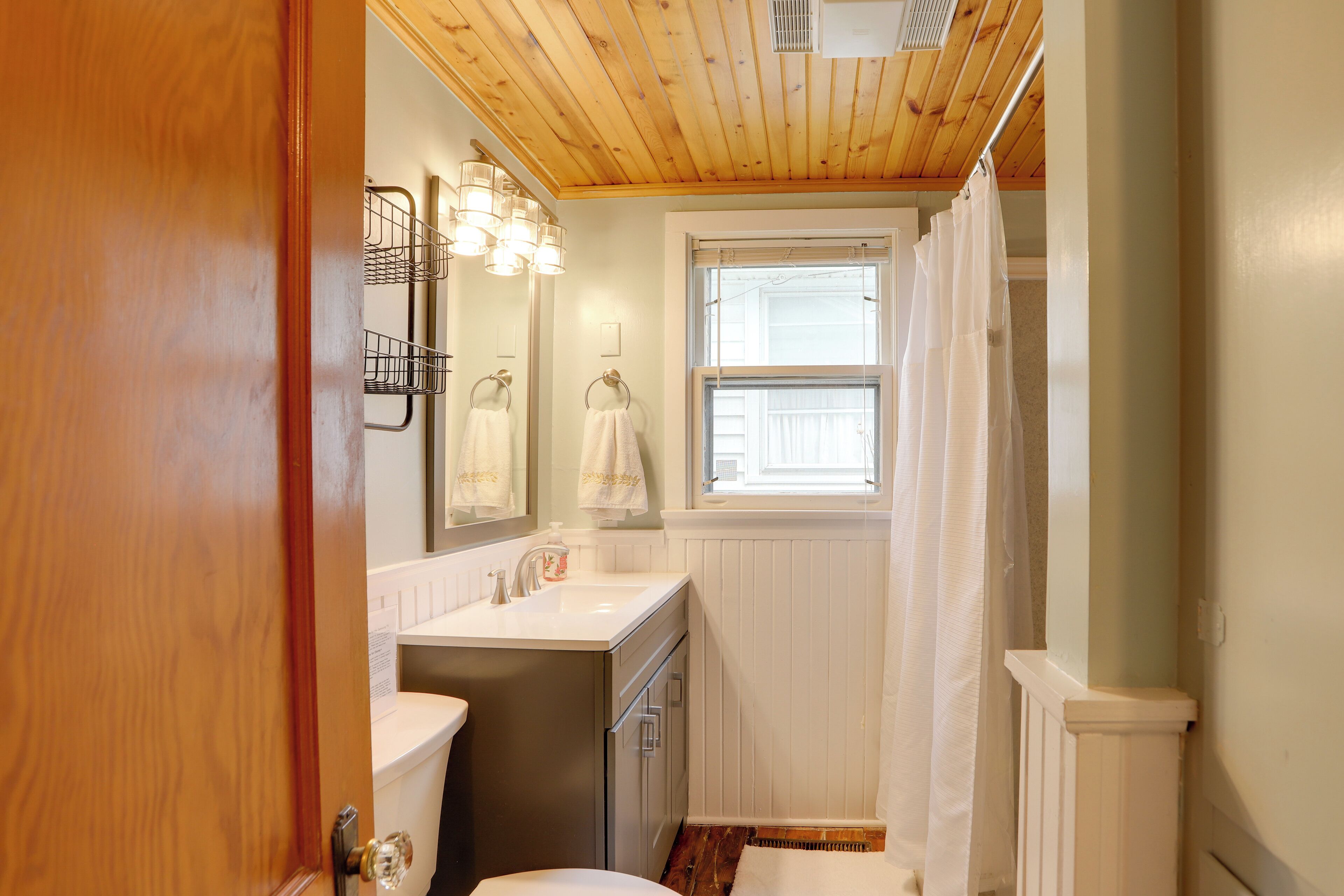 A compact bathroom with a wooden ceiling, a white toilet, a sink with a mirror above it, and a window with a white curtain.