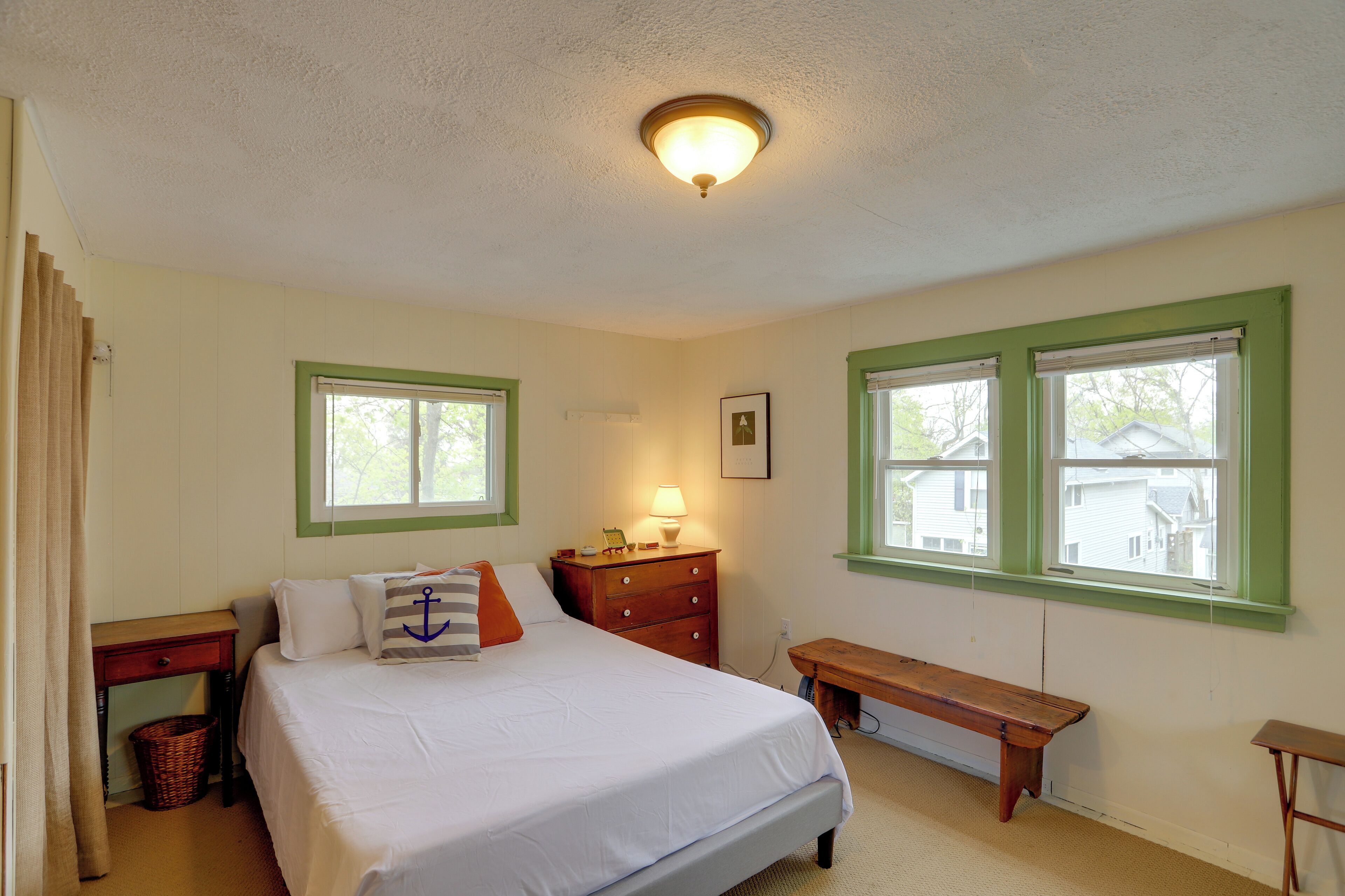 A bedroom with a bed, a nightstand, a dresser, a bench, and two windows with green trim.