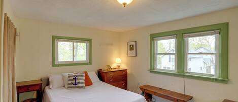 A bedroom with a bed, a nightstand, a dresser, a bench, and two windows with green trim.