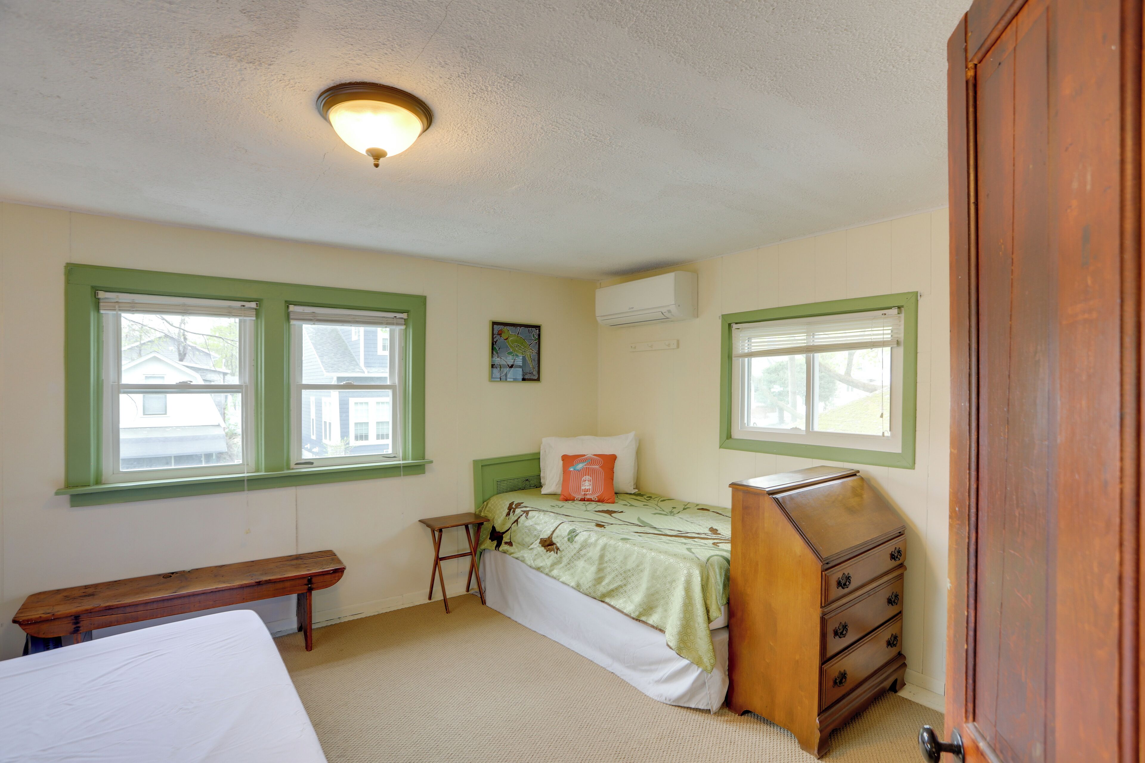 A bedroom with a bed, a bench, a dresser, and two windows with green frames.