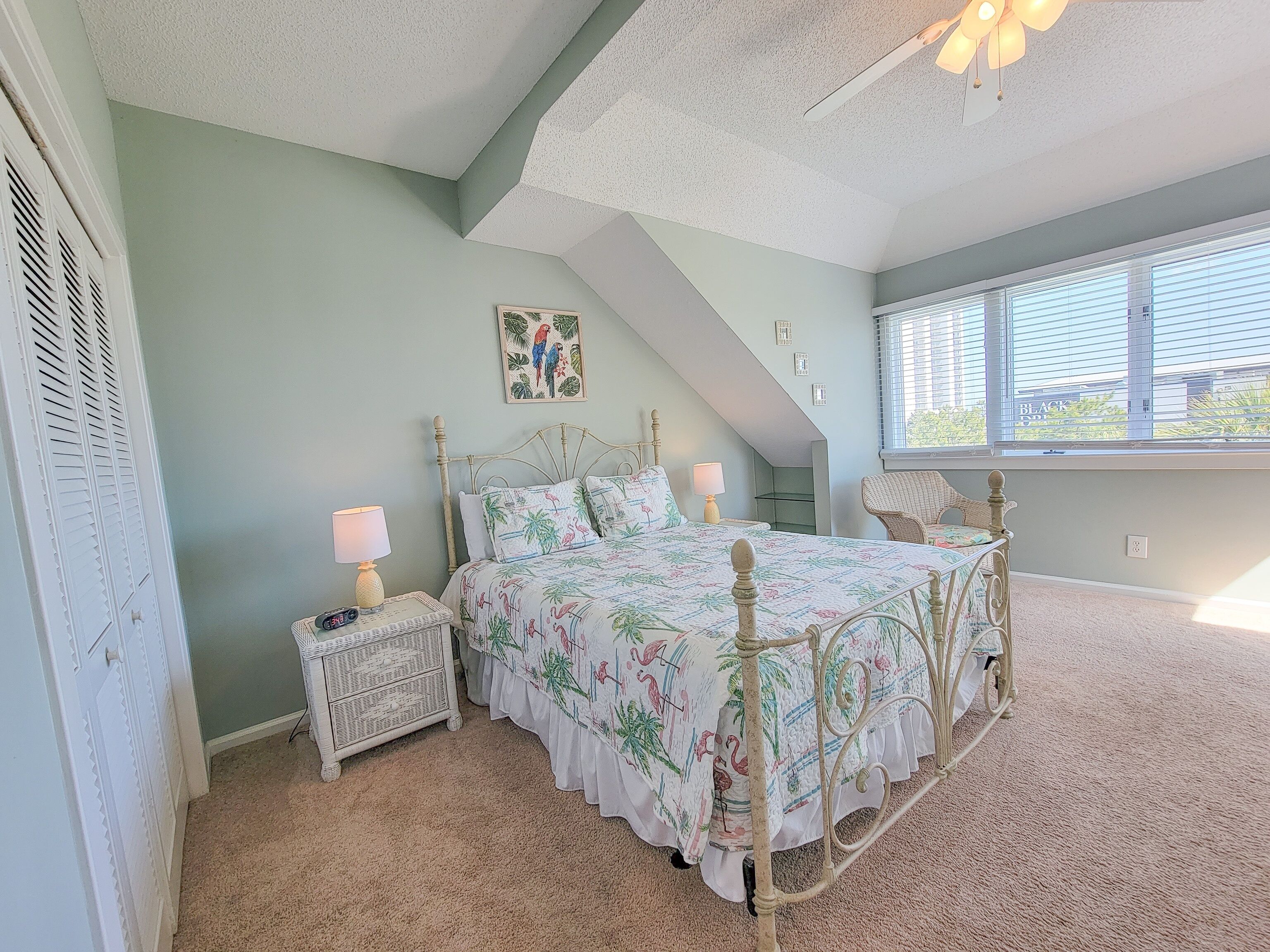 A bedroom with a bed, nightstand, and chair. There is a window with blinds and a ceiling fan.