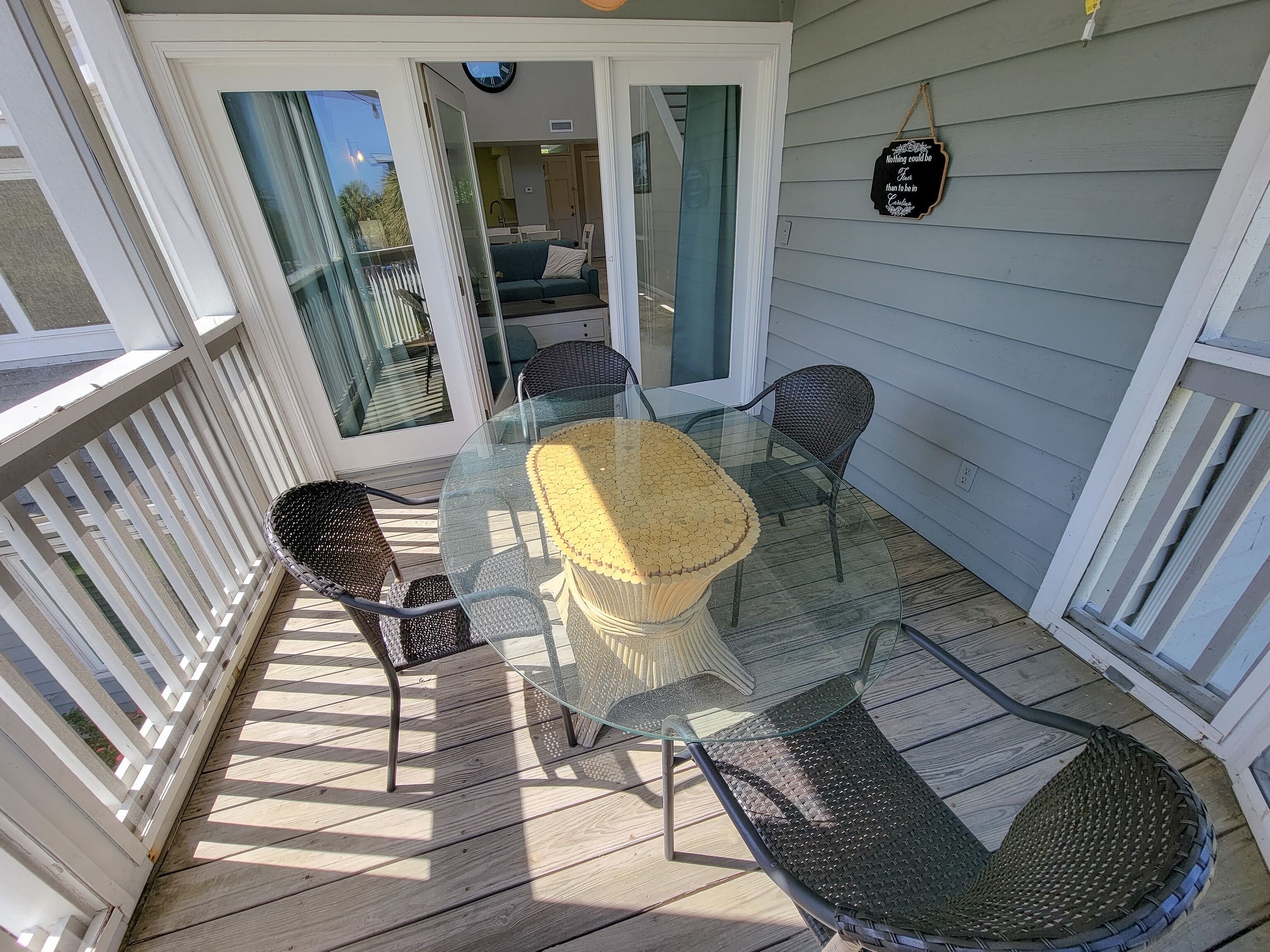Une terrasse couverte avec une table et des chaises en verre, donnant sur l’intérieur de la maison, et un panneau décoratif accroché au mur.