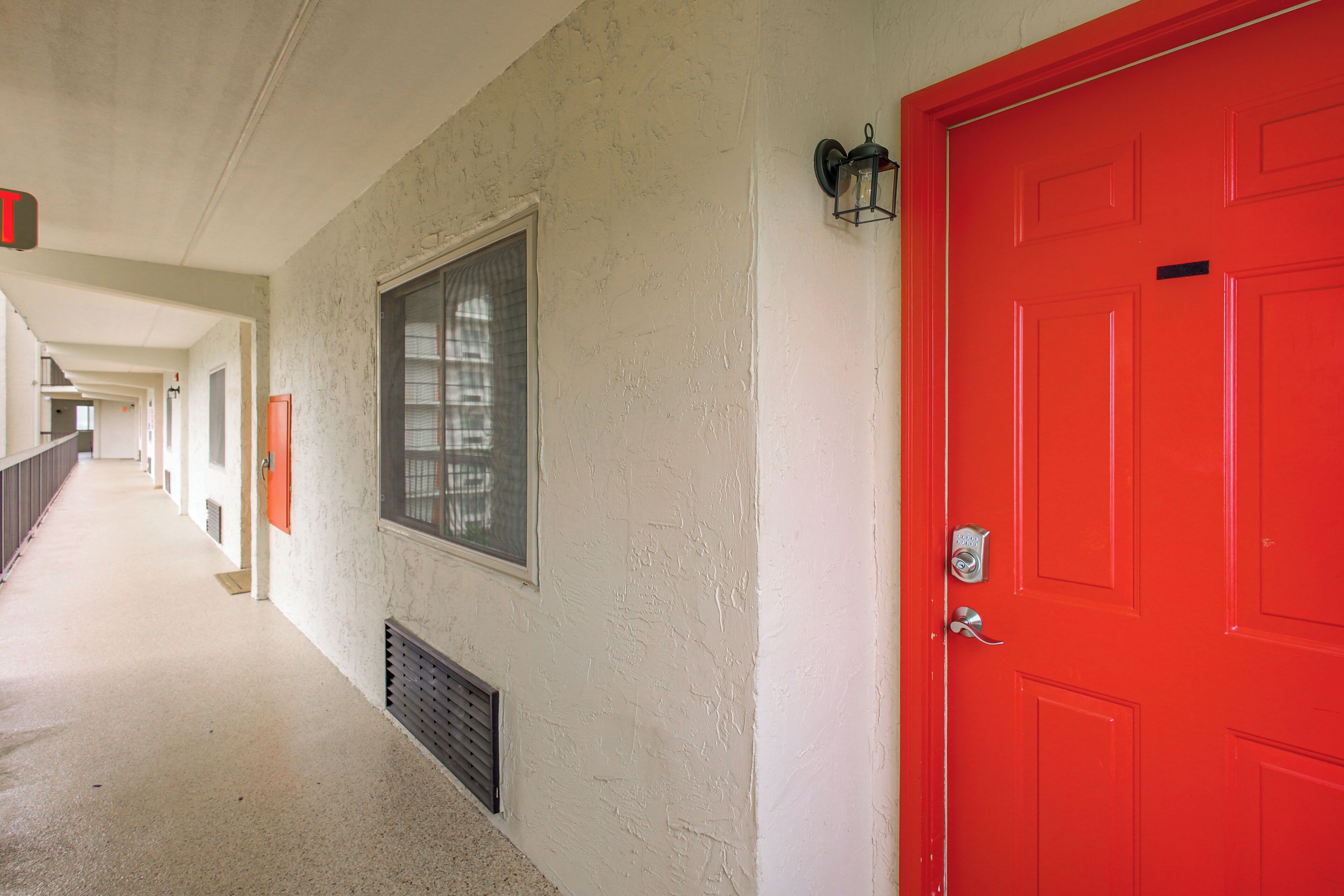 A corridor with a red door, a window, and a vent on the wall.