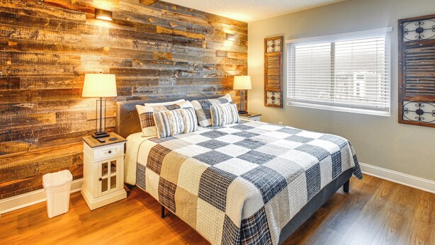 A bedroom with a wooden wall, a large bed with a checkered quilt, two bedside tables with lamps, and a window with blinds. - Gatlinburg Condo w/ Smoky Views: Walk to Town! (Gatlinburg)