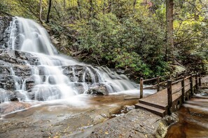 Ein Wasserfall stürzt über Felsen, eine Holzbrücke führt zu einer Steinplattform.