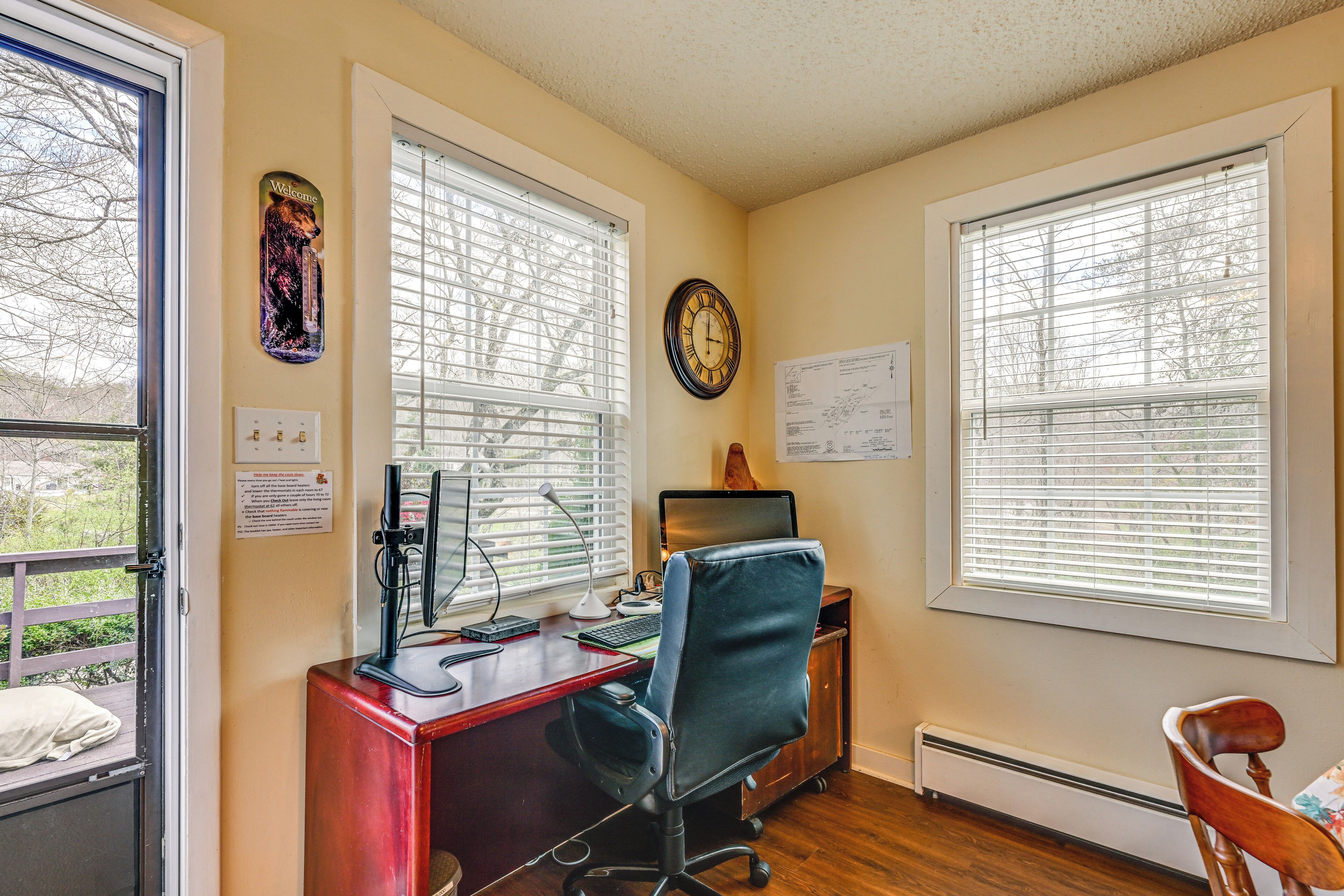 A home office with a desk, computer, and chair, large windows with blinds, and a clock on the wall.