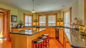 A kitchen with wooden cabinets, a central island with stools, a sink, and a window with blinds. - Historic Manteo House on Roanoke Sound w/ Dock! (Manteo)