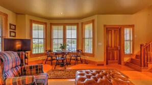 A spacious living room with a large brown leather ottoman, plaid armchairs, a dining table with chairs, and a television. - Historic Manteo House on Roanoke Sound w/ Dock! (Manteo)
