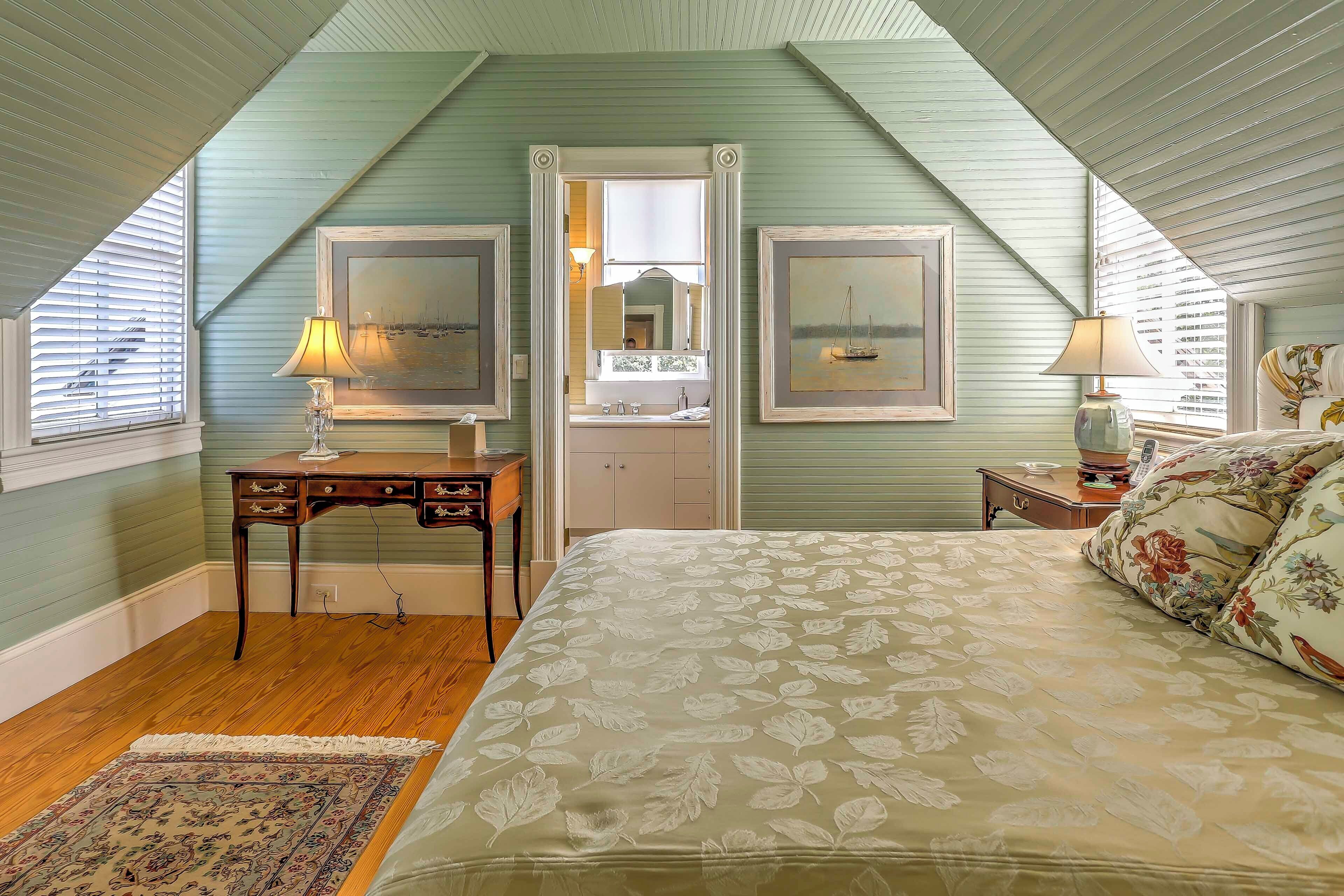 A bedroom with a sloped ceiling, a bed with floral bedding, a desk with a lamp, and framed artwork on the walls.