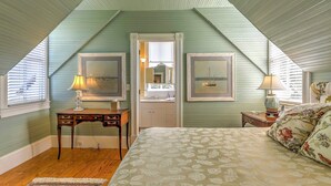 A bedroom with a sloped ceiling, a bed with floral bedding, a desk with a lamp, and framed artwork on the walls. - Historic Manteo House on Roanoke Sound w/ Dock! (Manteo)