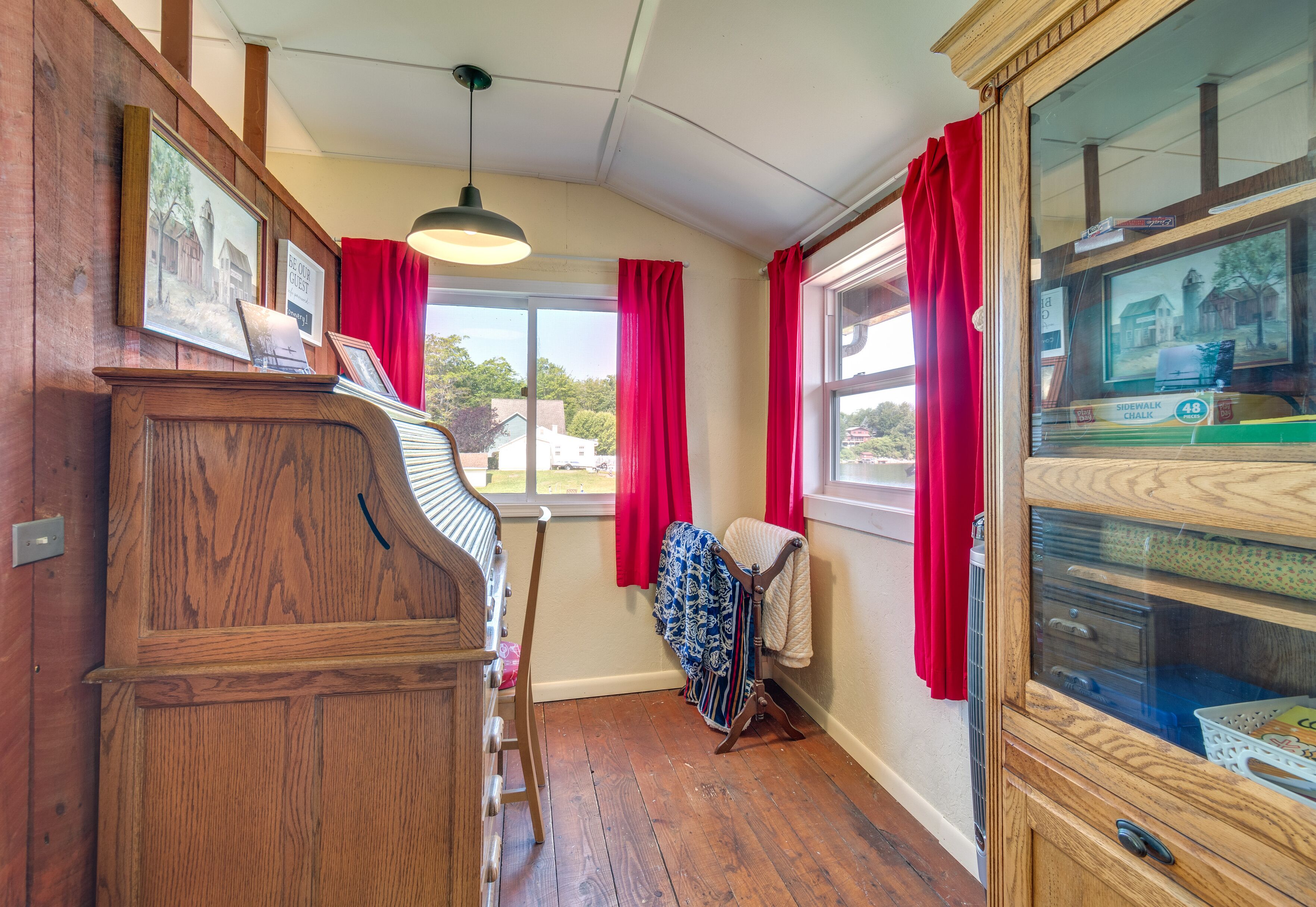 Quarto com piano de madeira, janela com cortinas vermelhas, luminária pendente e armário de madeira com gavetas.