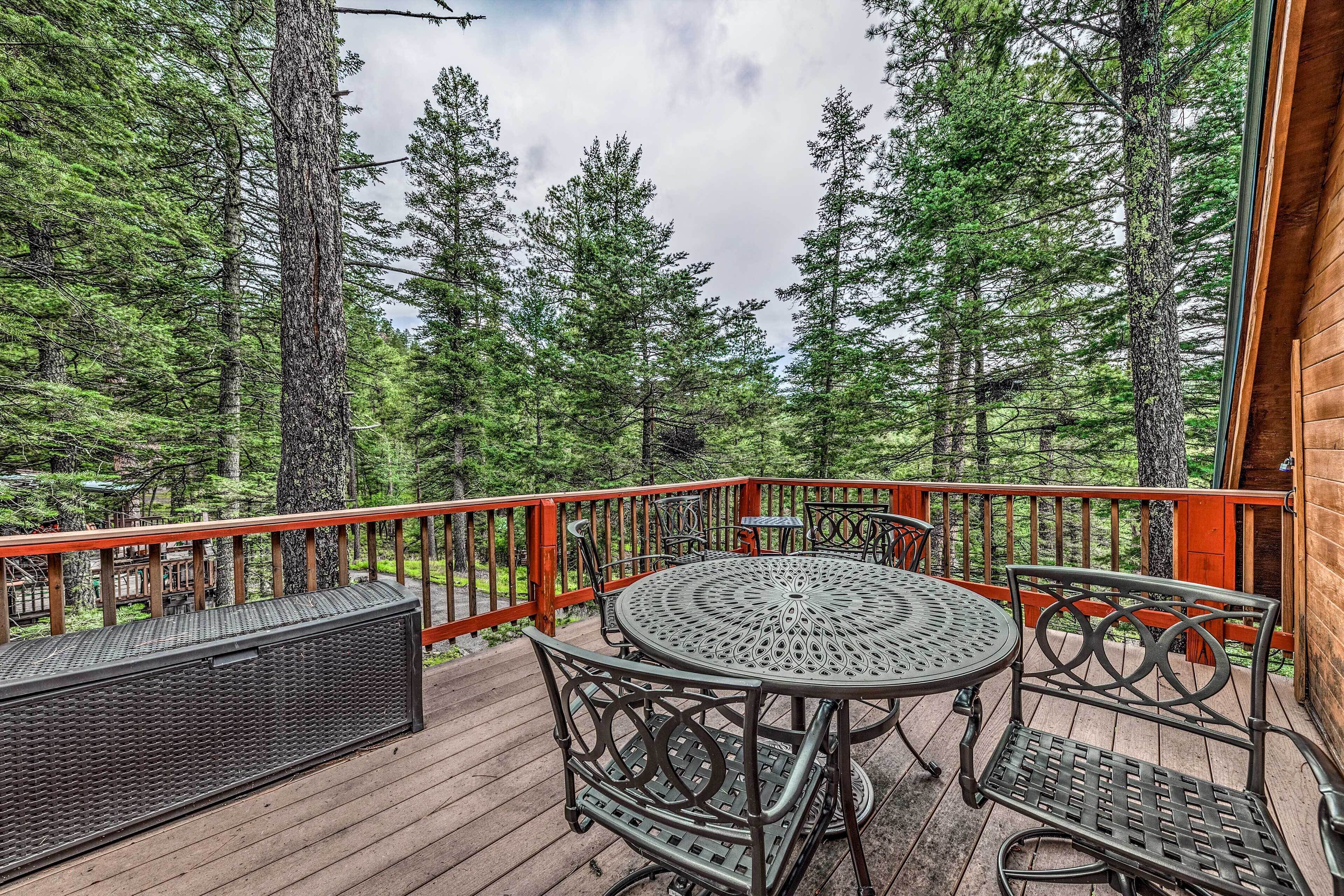 A wooden deck with a round table and two chairs, surrounded by tall trees.