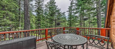 A wooden deck with a round table and two chairs, surrounded by tall trees.
