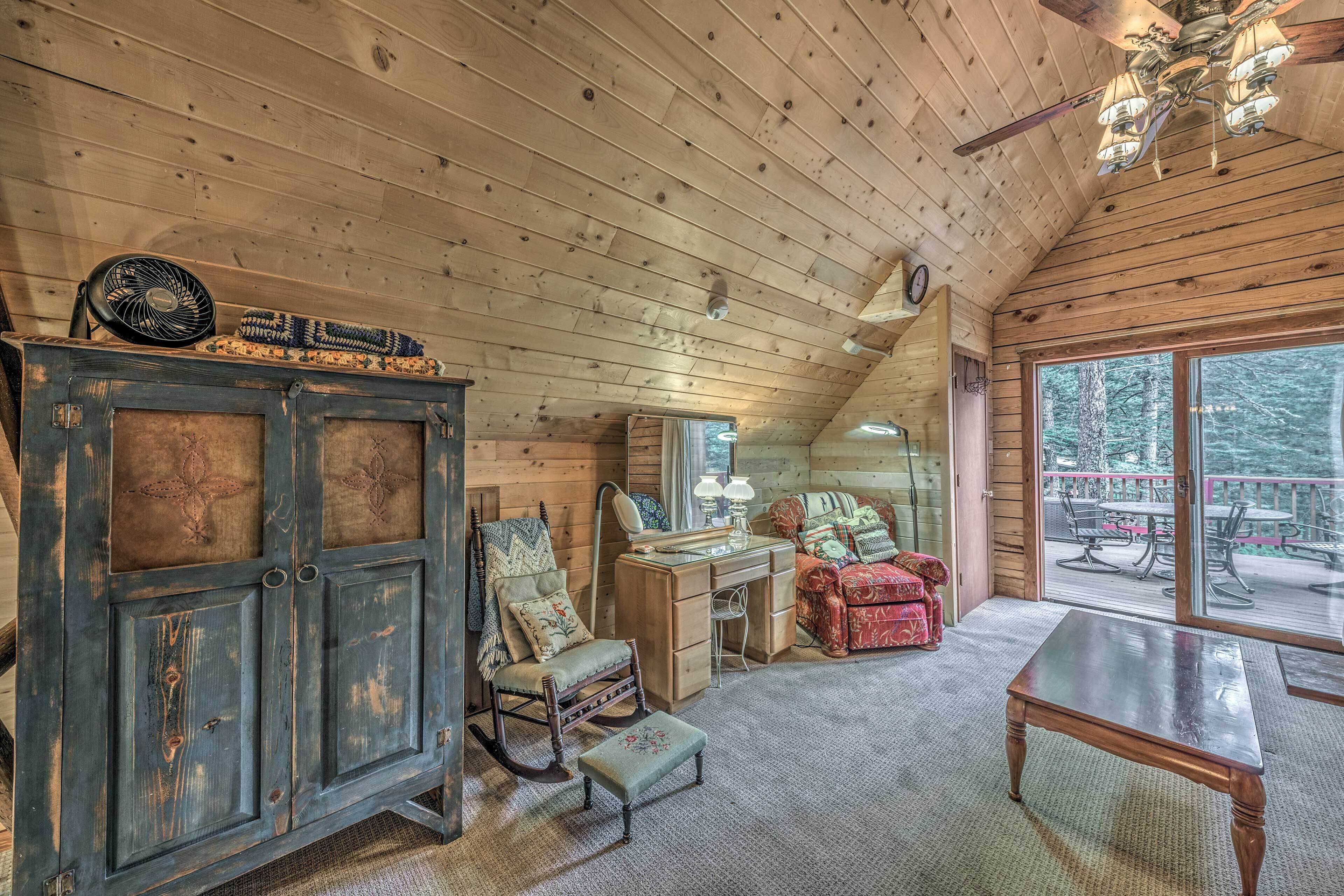 A cozy cabin interior with a wooden ceiling, a rocking chair, a desk, and a large window overlooking a deck.