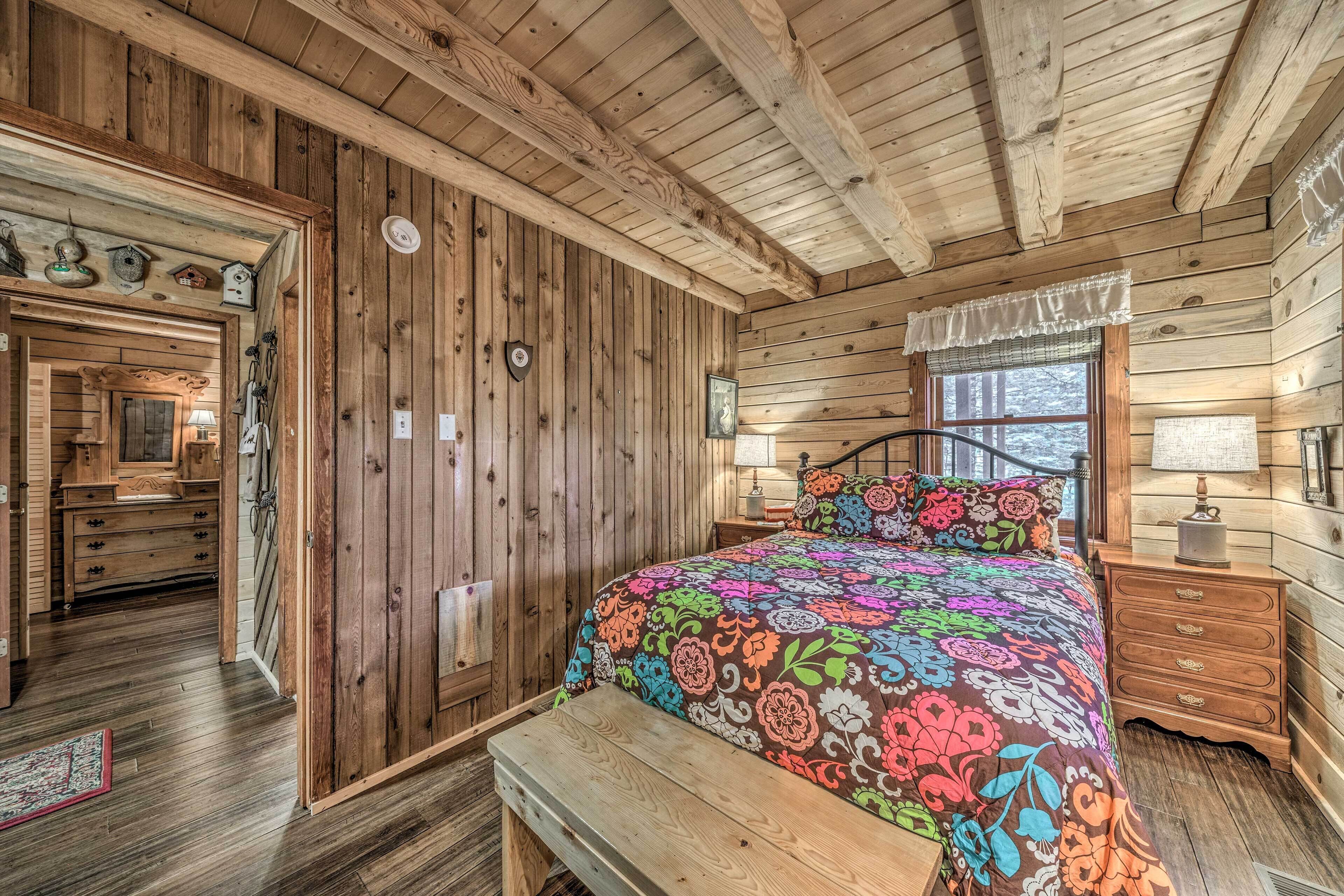 A wooden cabin bedroom with a bed, bench, dresser, and a window with a view.