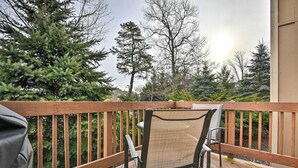 A wooden deck with a chair and a grill, surrounded by trees and a house in the background.