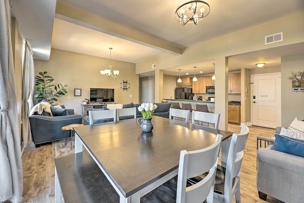 A spacious living room with a dark wooden dining table, white chairs, and a chandelier. There is a sofa with a vase of flowers and a potted plant. The kitchen is visible in the background with modern appliances.