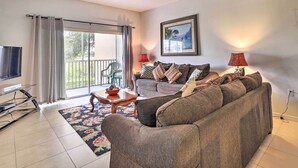A living room with a sofa, a TV, a coffee table, and a balcony with a view of greenery.
