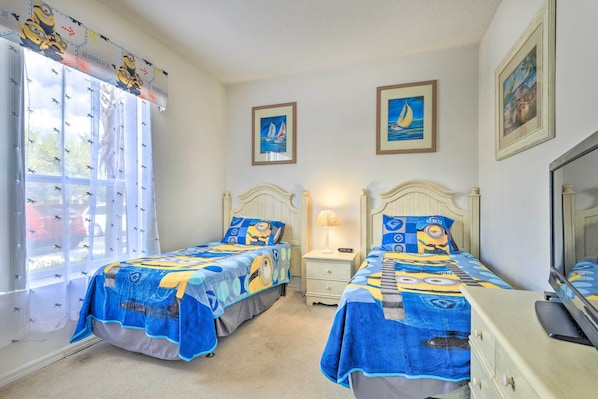 Une chambre avec deux lits, chacun recouvert d’une couverture bleue ornée d’un personnage de dessin animé. Deux tableaux encadrés sont accrochés au mur, il y a une fenêtre avec des rideaux et une table de chevet avec une lampe placée entre les deux lits.