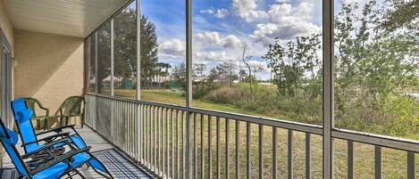 A covered outdoor area with a view of a grassy landscape and trees. There are two blue chairs and a small table.