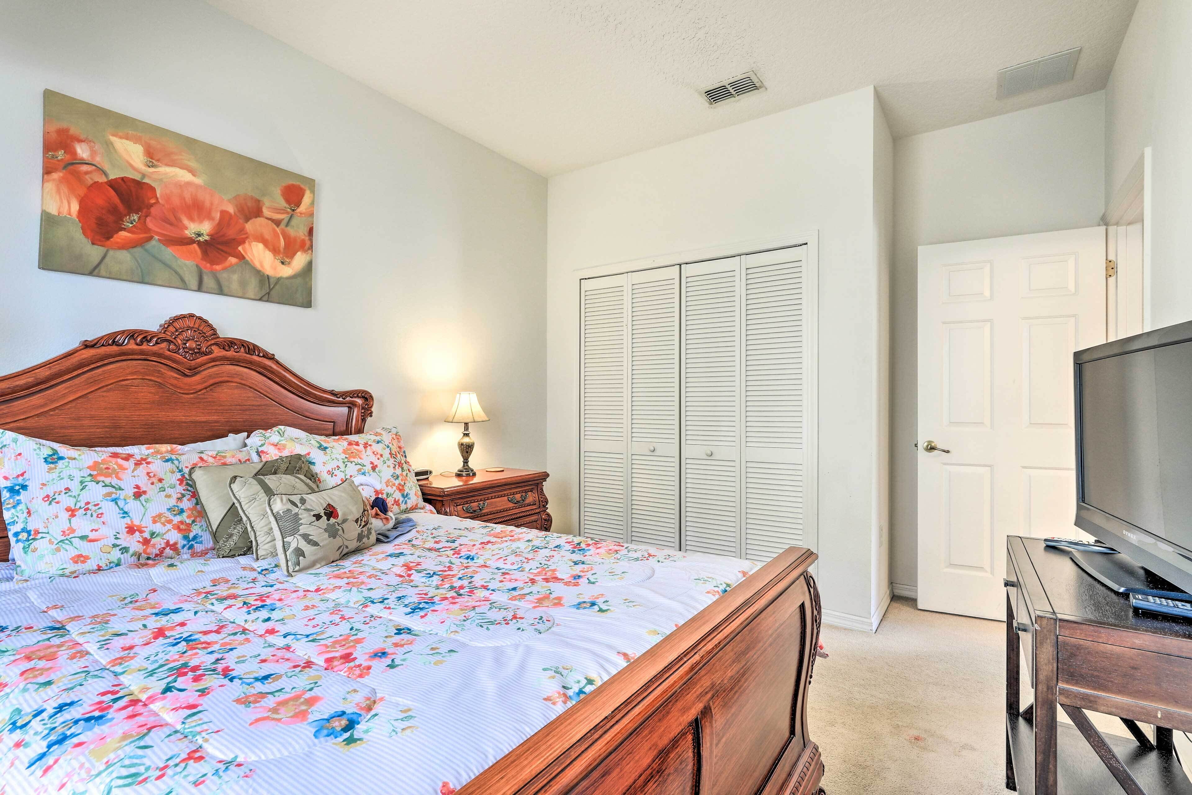 Un dormitorio con cama de madera, ropa de cama floral, un cuadro en la pared, una lámpara de noche, un armario y un televisor sobre un mueble de madera.