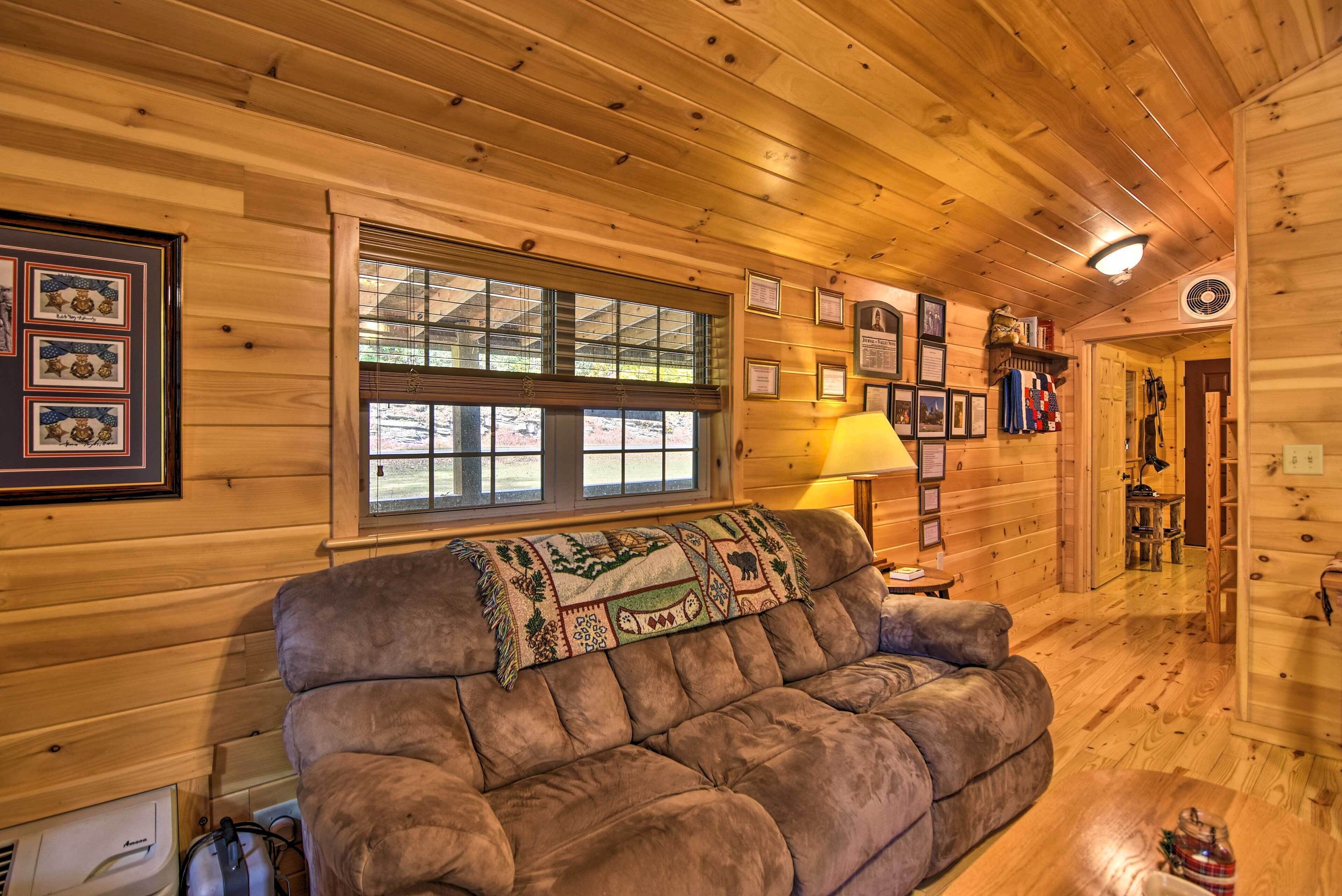 A cozy log cabin interior with a plush sofa, framed pictures, a window with blinds, and a lamp.