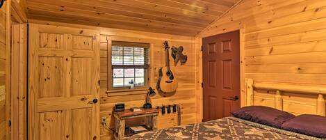 A wooden cabin bedroom with a bed, a desk, a guitar, and a window.