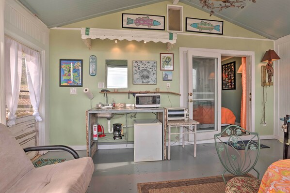 A compact kitchen with a microwave, toaster, and refrigerator. There is a small dining area with a chair and a table. The room has a door leading to another space and a window with curtains. - Lakeside Pleasure Island Cabin w/ Deck & Gas Grill (Farmerville)