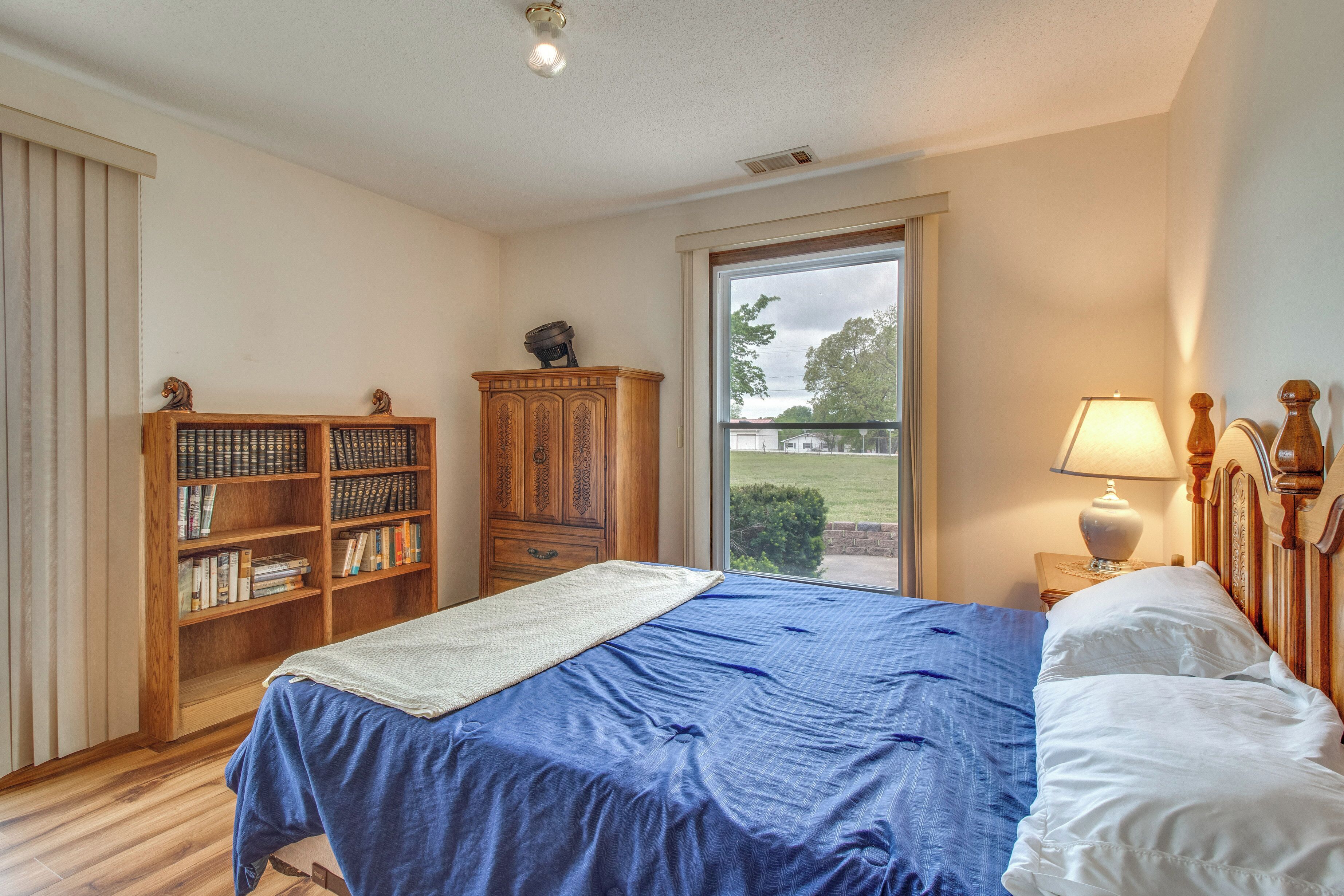 A bedroom with a large bed, a wooden bookshelf, a window with a view, and a nightstand with a lamp.
