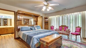 A bedroom with a large bed, a wooden headboard, a nightstand, a dresser, a vanity with a mirror, a ceiling fan, and a window with blinds. - Gassville Home 3 Mi to White River & Trout Fishing (Gassville)