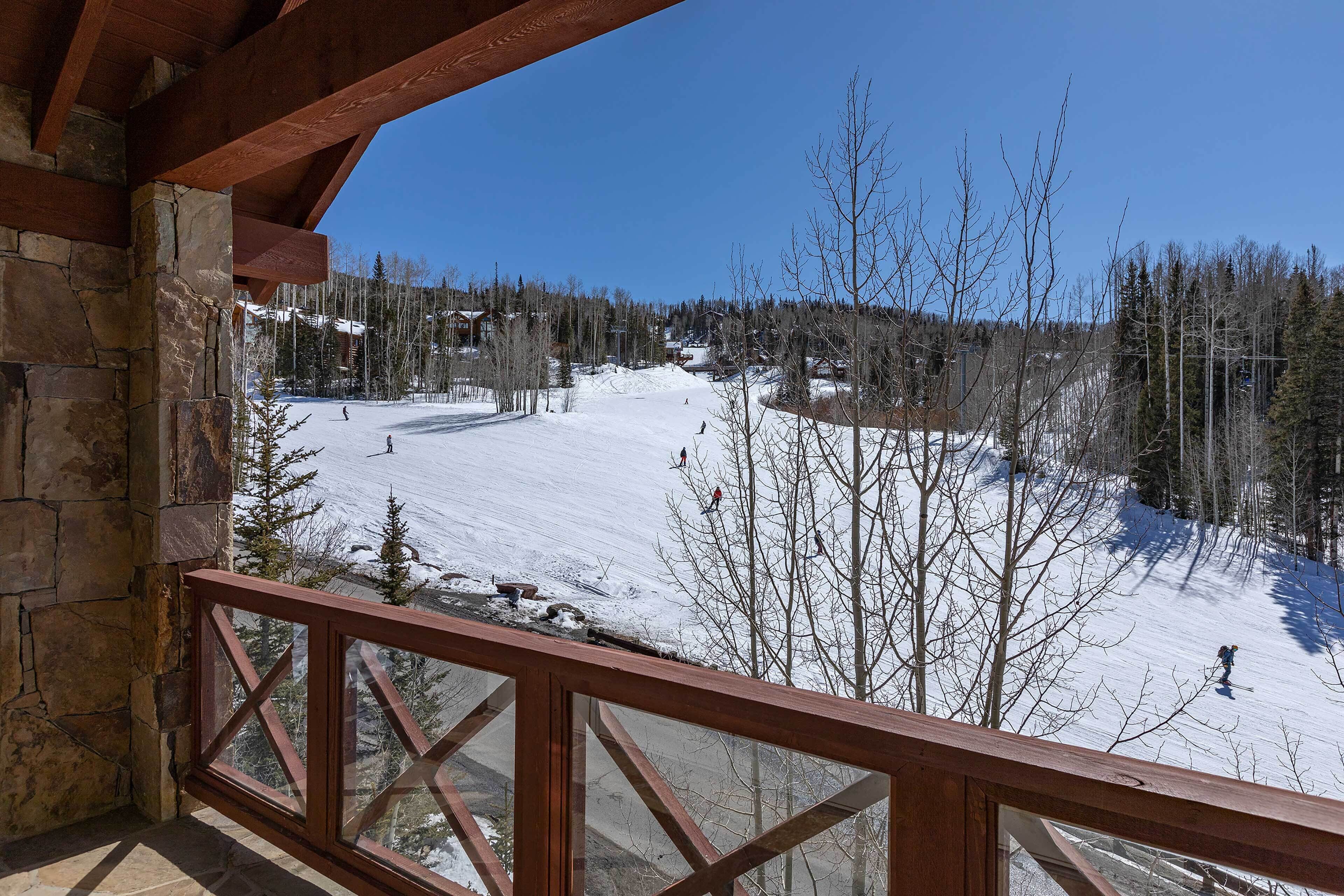 A snow-covered ski slope with skiers, a stone building with a wooden balcony, and a clear blue sky.