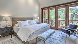 Un dormitorio con una cama grande, mesitas de noche, un banco y vistas a un paisaje montañoso a través de ventanas amplias.