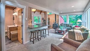 A spacious living area with a kitchen, bar stools, and a large window providing a view of the outdoors. - Peace in the Pines: Cle Elum Cabin w/ Trail! (Cle Elum)
