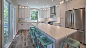 Une cuisine moderne avec des meubles blancs, un grand îlot central doté de tabourets, des appareils en acier inoxydable et une vue sur la verdure à l’extérieur.