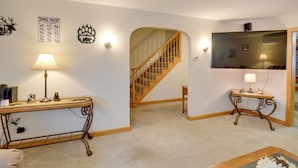 Una sala de estar con escalera de madera, televisor de pantalla plana, lámpara y una mesa con figuritas.