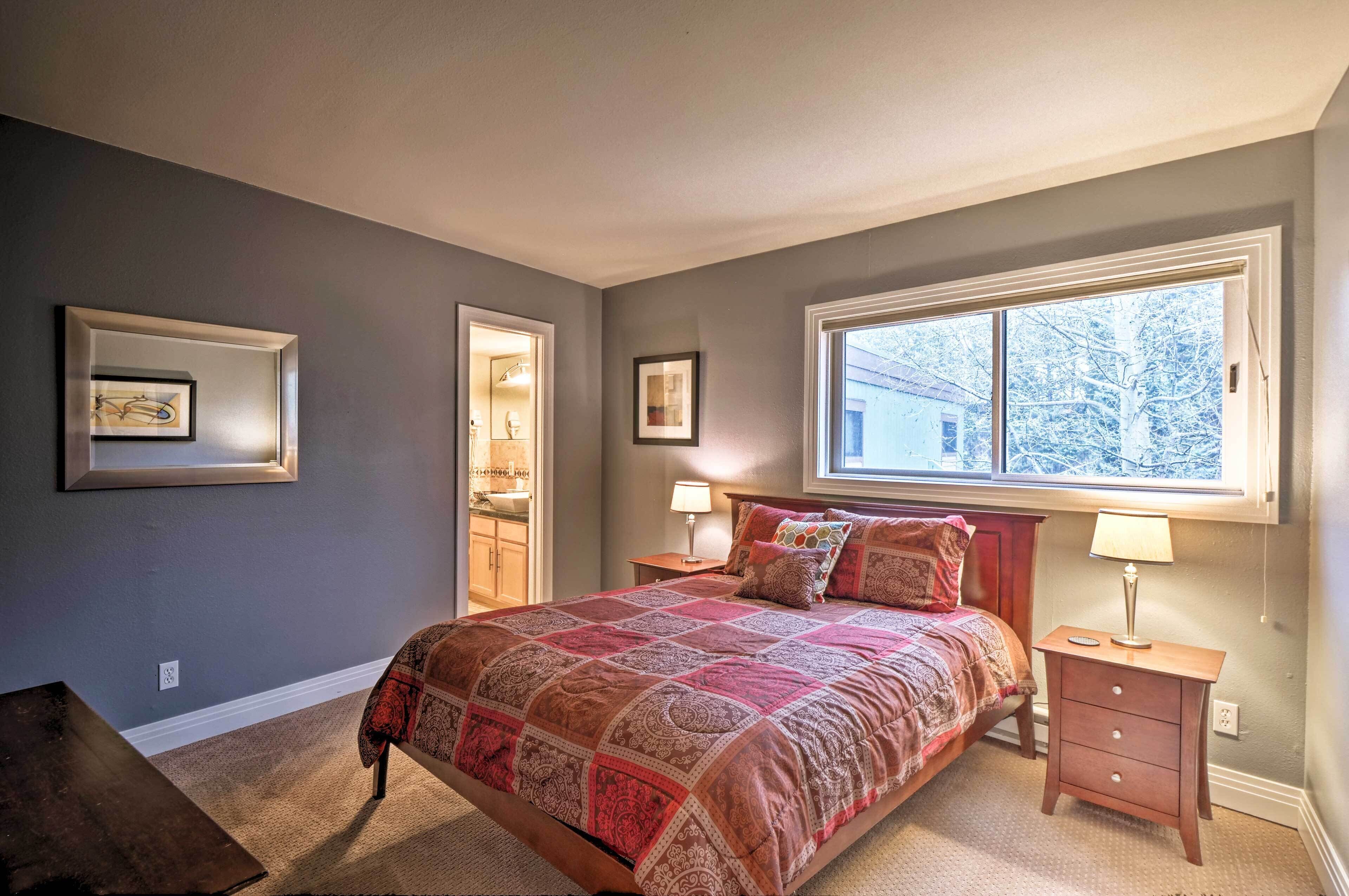 A bedroom with a large bed, two bedside tables with lamps, a window with a view of trees, and framed pictures on the wall.