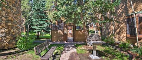 A stone building with a wooden door, a bench, and a grill area in a garden with trees and a paved path.