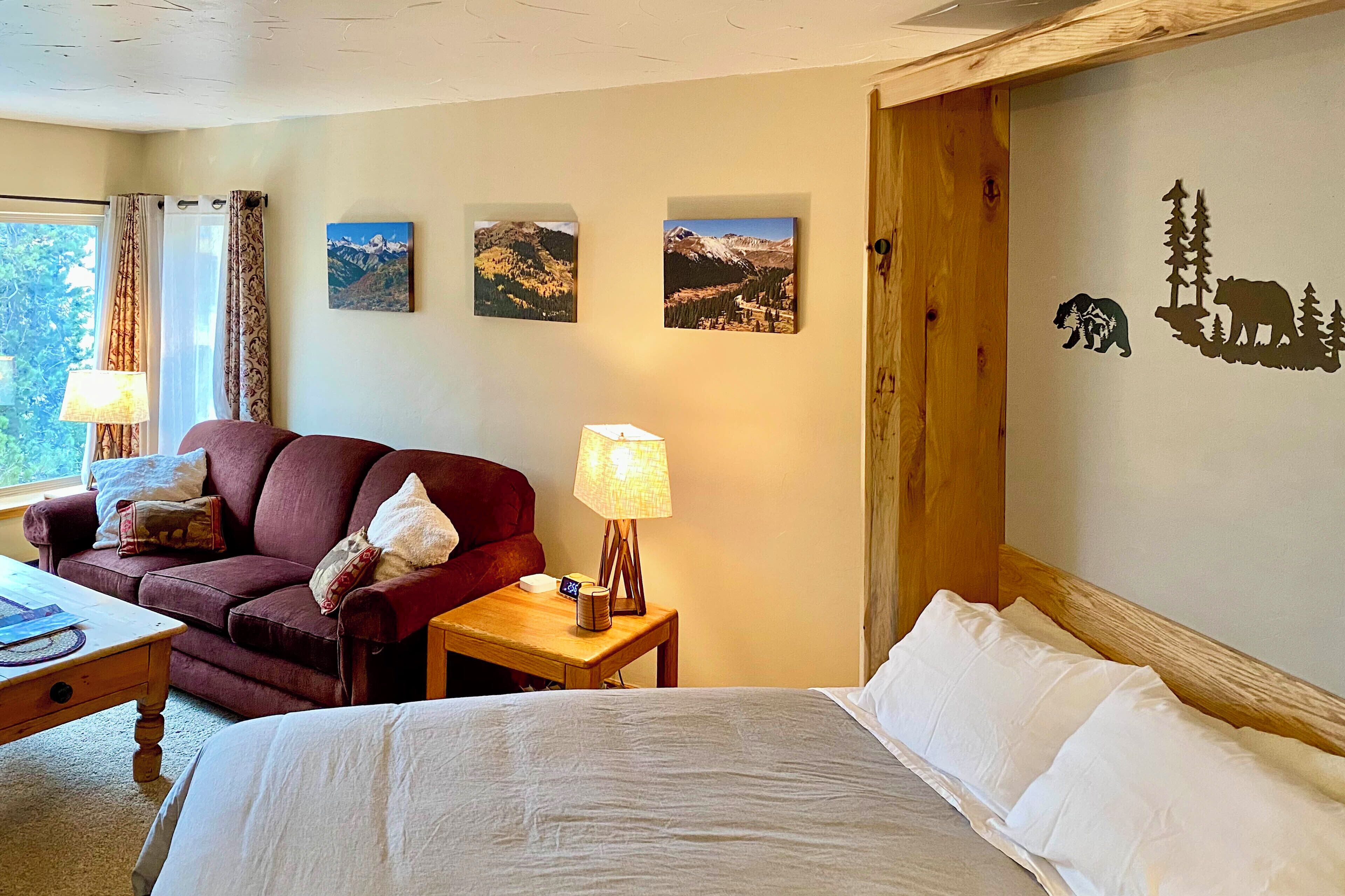 A hotel room with a bed, a wooden nightstand with a lamp, a red sofa, and two wall-mounted pictures of mountains.