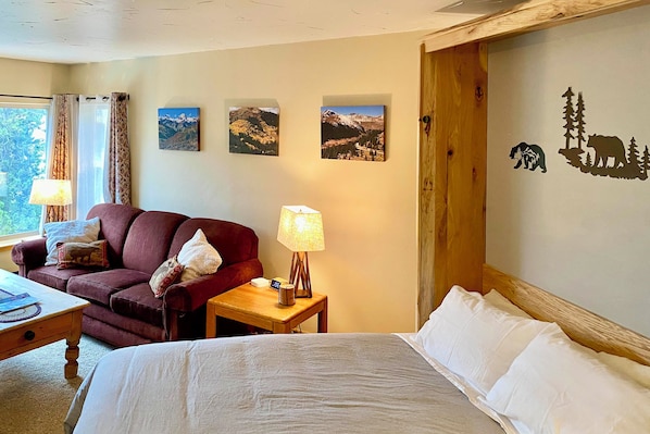 Una habitación de hotel con una cama, una mesita de noche de madera con una lámpara, un sofá rojo y dos cuadros de montañas colgados en la pared.