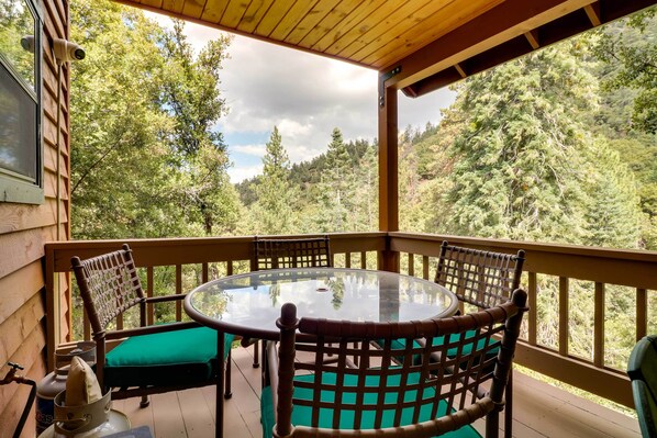 Una terraza de madera con una mesa redonda de vidrio y sillas, con vistas a una ladera boscosa.