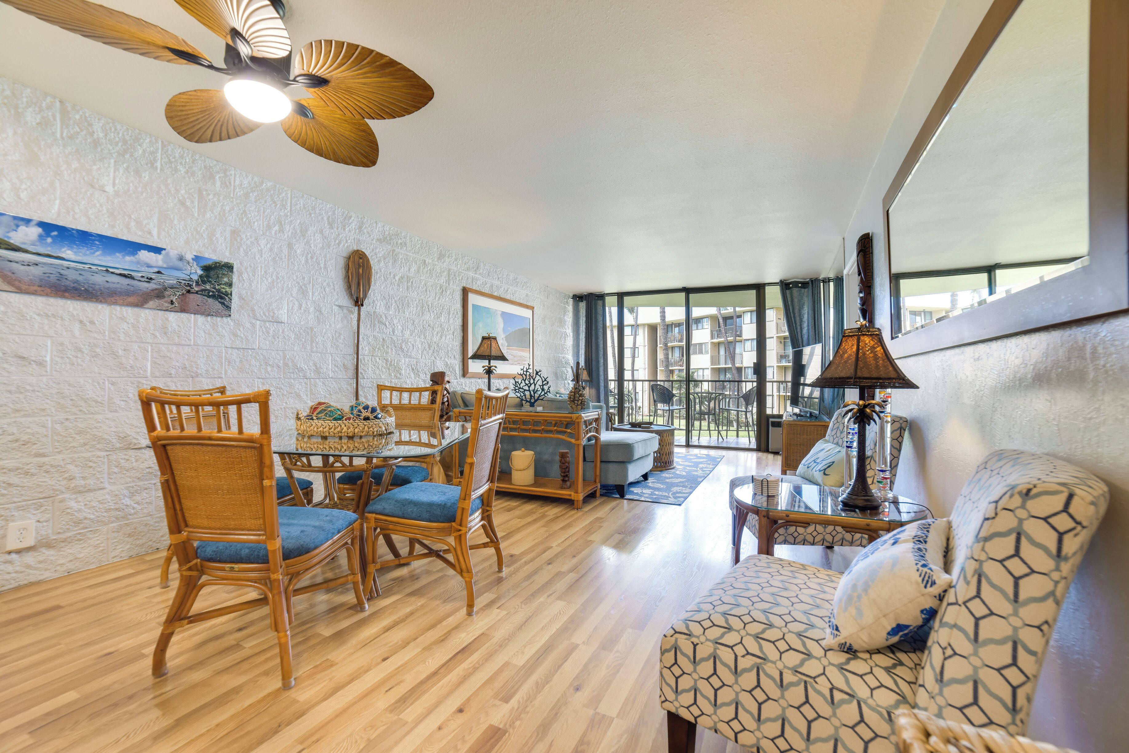 A spacious living room with a wooden floor, a dining area with wicker chairs, a patterned sofa, and a ceiling fan.
