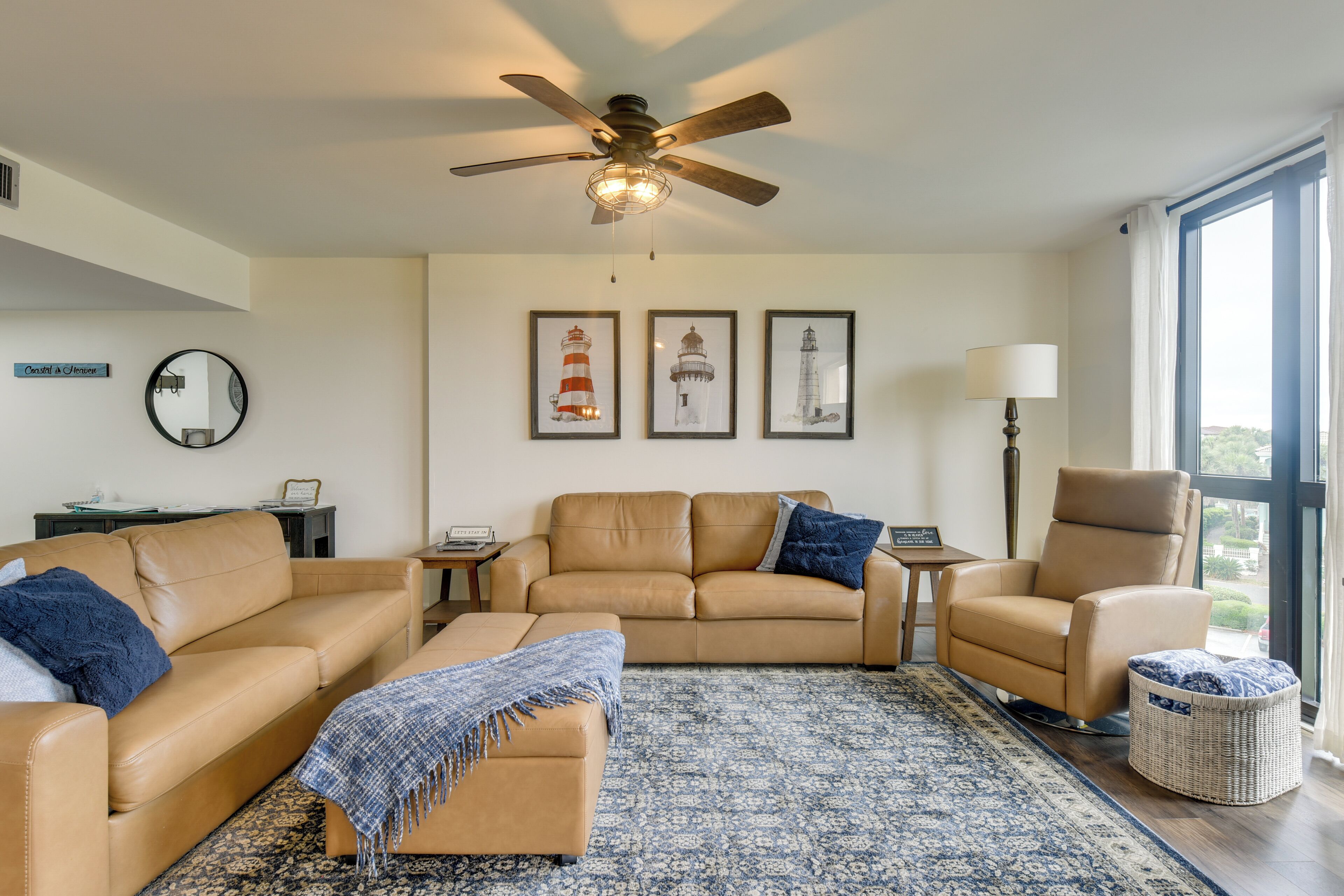 Un salon avec un ventilateur de plafond, un canapé, un fauteuil, une petite table et une lampe. Trois tableaux encadrés sont accrochés au mur.