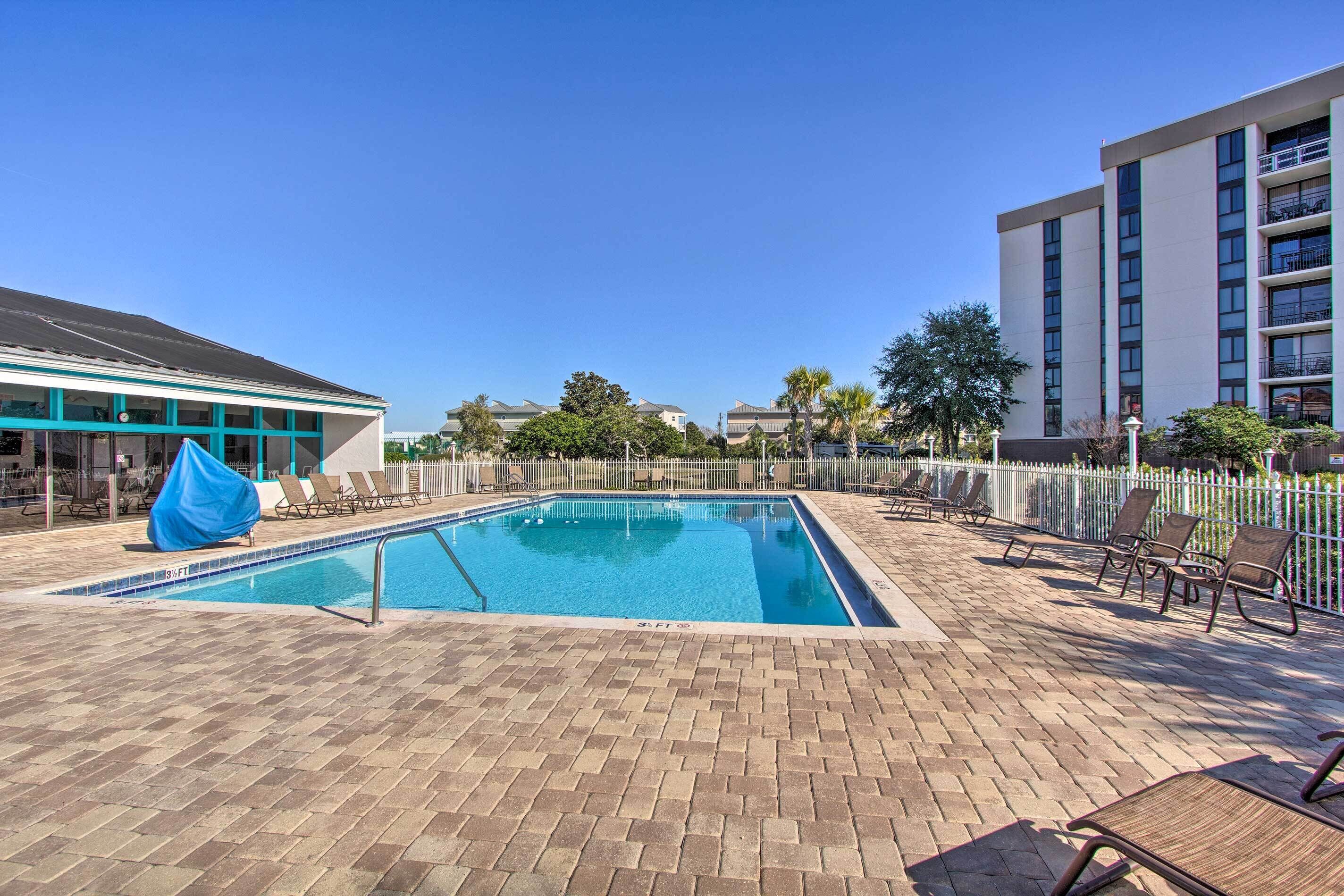 A swimming pool with lounge chairs and a covered area, surrounded by a brick patio and a white fence.