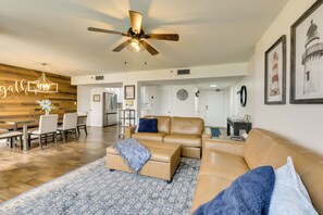 A spacious living room with a brown leather sofa, a blue patterned rug, a ceiling fan, and a dining area with a wooden wall and a neon sign. - Airy Destin Condo w/ Pool - Walk to Private Beach! (Destin)