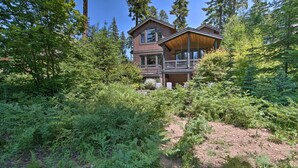 A two-story log cabin with a balcony, surrounded by dense forest. - Modern Alpine Home on Prospector Golf Course! (Cle Elum)